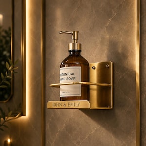 Peut inclure: Un support mural en laiton pour distributeur de savon avec une étagère rectangulaire, une barre de protection et l'inscription gravée JOHN & EMILY. Il contient une bouteille en verre brun avec une pompe et une étiquette BOTONICAL HAND SOAP.