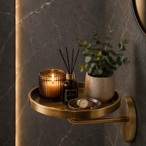 Puede incluir: Un estante de pared circular de latón que sostiene una vela de vidrio ámbar estriado con llama encendida, un frasco difusor de vidrio oscuro, una pequeña maceta de cerámica con una planta verde y un plato con un reloj de pulsera dorado.