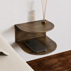 May include: A wall-mounted, two-tiered floating shelf with a dark, brushed bronze finish. The shelf features a semi-circular design with two levels. A small reed diffuser sits on the top shelf, and a dark smartphone rests on the bottom shelf.