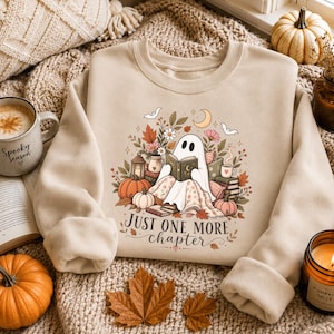 Könnte beinhalten: Ein beigefarbenes Sweatshirt mit Rundhalsausschnitt und einer Grafik eines geisterhaften Wesens, das ein Buch liest, umgeben von Kürbissen, Herbstblättern, Blumen und Fledermäusen. Der Text "JUST ONE MORE chapter" steht in brauner Schrift darunter.