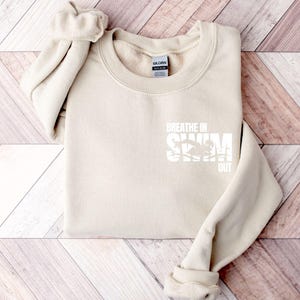 Könnte beinhalten: Ein sandfarbenes Sweatshirt mit Rundhalsausschnitt und einem weißen Aufdruck auf der Brust. Der Aufdruck zeigt den Text BREATHE IN SWIM OUT mit der Silhouette eines Schwimmers in der Mitte. Das Kleidungsstück hat lange Ärmel sowie gerippte Bündchen.