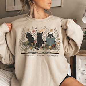 Könnte beinhalten: Ein beigefarbenes Sweatshirt mit Rundhalsausschnitt und einer grafischen Illustration von drei schwarzen Katzen zwischen Wildblumen und einem offenen Buch. Eine Katze liest, eine wird gestreichelt, eine versteckt sich hinter einem Buch. Text: READ BOOKS + PET CATS + AVOID PEOPLE.