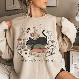 Könnte beinhalten: Ein beigefarbenes Sweatshirt mit Rundhalsausschnitt und einem Grafikdruck einer schwarzen Katze, die auf einem Stapel aus drei Büchern schläft. Neben den Büchern steht eine weiße Kaffeetasse mit einem Herzsymbol. Das Design zeigt bunte Wildblumen, einen Schmetterling und den Schriftzug "cats • coffee • chapters" in schwarzer Schreibschrift.