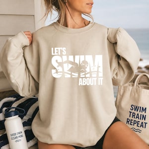 Könnte beinhalten: Ein beigefarbenes Sweatshirt mit Rundhalsausschnitt und weißem Aufdruck LET'S SWIM ABOUT IT, ergänzt durch eine Grafik eines Schwimmers in Bewegung. Das Sweatshirt hat lange Ärmel mit gerafften Schultern und eine lockere Passform.