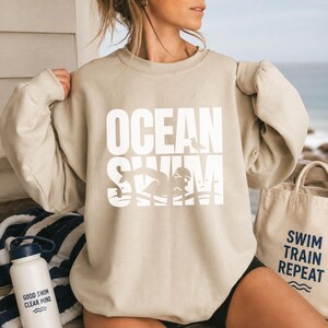 Könnte beinhalten: Ein beigefarbenes Sweatshirt mit Rundhalsausschnitt und dem weißen Schriftzug "OCEAN SWIM", einer Grafik einer schwimmenden Person und einem kleinen Vogel. Das Sweatshirt hat lange Ärmel mit gerafften Schultern. Eine weiße Trinkflasche ist zu sehen.