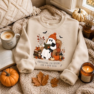 Könnte beinhalten: Ein beigefarbenes Sweatshirt mit Rundhalsausschnitt und einer Grafik eines Geistes mit orangefarbener Mütze, der in einem Sessel sitzt und mit einer schwarzen Katze ein Buch liest. Der Text lautet GHOSTING EVERYONE FOR A GOOD BOOK, umgeben von Kürbissen.