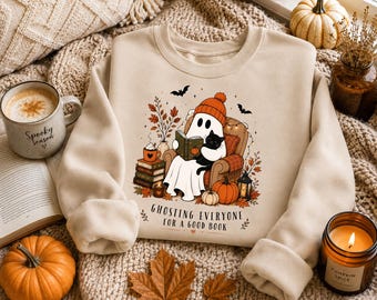 May include: A beige crewneck sweatshirt featuring a graphic of a ghost wearing an orange beanie, sitting in an armchair reading a book with a black cat. The design includes pumpkins, autumn leaves, and the text Ghosting everyone for a good book.