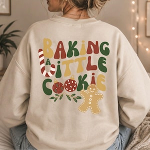 Könnte beinhalten: Ein beiges Sweatshirt mit Rundhalsausschnitt und einem großen Aufdruck auf der Rückseite mit dem Text "BAKING A LITTLE COOKIE" in wellenförmigen, bunten Buchstaben. Das Design zeigt eine Zuckerstange, zwei rote Weihnachtskugeln, Stechpalmen und einen Lebkuchenmann.