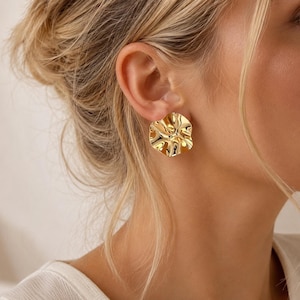 Peut inclure: Une boucle d'oreille à clou dorée présentant une texture de surface circulaire, irrégulière et ondulée. La boucle d'oreille possède une finition polie et réfléchissante qui capte la lumière, créant un effet de vague sur la surface métallique.