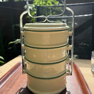 May include: A four-tiered stackable food container with a pale yellow enamel finish and dark green rim accents. The cylindrical unit features a metal locking frame with a cylindrical wooden handle at the top for carrying the stacked compartments.