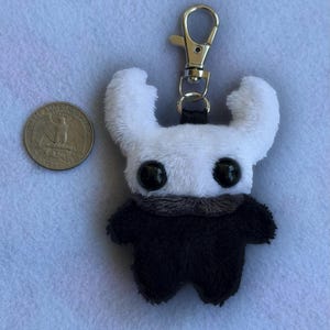 Puede incluir: Un llavero de peluche con cabeza blanca, dos grandes ojos negros y cuernos curvados, unido a un mosquetón de metal plateado. Una moneda de 25 centavos de EE. UU. está colocada al lado para comparar el tamaño, midiendo 24,26 mm de diámetro.