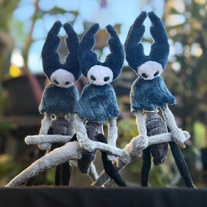 Puede incluir: Tres figuras de peluche inspiradas en el personaje Hollow Knight, cada una con una máscara blanca con cuencas negras, una capa con capucha azul oscuro y extremidades largas y delgadas. Las figuras están hechas de tela suave y agrupadas.