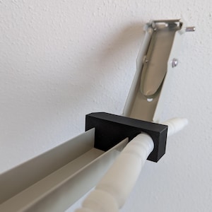 Op de afbeelding: Een witte metalen beugelconstructie met een zwart rechthoekig verbindingsblok. De structuur bevat een lichtgrijze schuine steunarm en een witte geribbelde cilindrische staaf die horizontaal door het zwarte verbindingsstuk steekt.