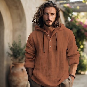 Puede incluir: Una camisa de manga larga con capucha en un color naranja quemado sólido. La prenda presenta una tapeta de botones parcial con tres botones visibles y cordones finos de color marrón oscuro que cuelgan del cuello de la capucha.