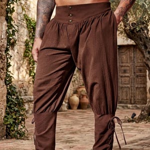 May include: Brown trousers featuring a high waistband with three metallic buttons, a loose-fitting pleated upper section, and fitted lower legs that include side lacing details with thin fabric ties at the ankles for a secure fit.
