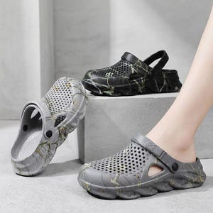May include: A pair of gray clogs and one black clog, each featuring a perforated upper, a pivoting heel strap, and a marbled pattern. The footwear has a thick, textured sole with side cutouts, providing a lightweight design for casual wear.