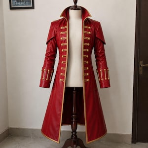 May include: A long, red coat featuring a high collar, structured shoulders, and gold-toned buttons arranged in vertical rows along the front opening and cuffs. The coat has gold piping along the edges and a flared hem, displayed on a mannequin.