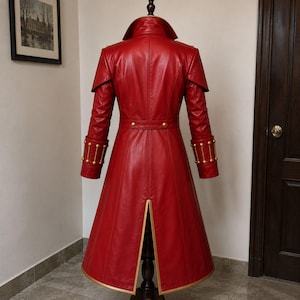 May include: A long red coat featuring a high collar, shoulder capes, and a waist belt with gold-toned buttons. The cuffs are adorned with vertical gold-toned bars and studs. The coat has a central back vent with gold trim along the bottom hem.