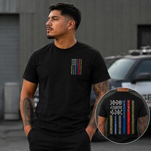 May include: A black short-sleeve t-shirt featuring a small graphic on the left chest and a large graphic on the back. Both graphics display vertical colored stripes representing emergency services with the text NO ONE FIGHTS ALONE.