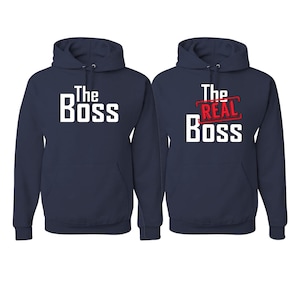 May include: Two navy blue hooded sweatshirts with long sleeves and front pockets. The left hoodie features the white text "The Boss". The right hoodie displays the white text "The Boss" with the red text "The Real" printed above it.