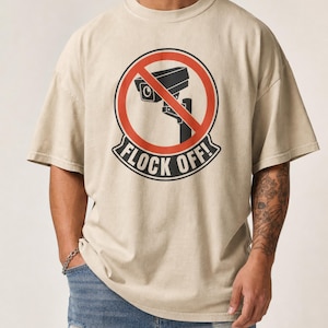 May include: A beige short-sleeved t-shirt featuring a graphic print of a black security camera inside a red prohibition circle. Below the circle, the text FLOCK OFF! is printed in bold black capital letters on a curved banner design.