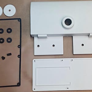 May include: A white metal enclosure kit consisting of a main housing with a central circular opening, a rectangular base plate, a thin black gasket frame, two white circular spacers, four small black washers, and two small black cylindrical fasteners.
