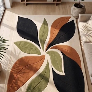 May include: A rectangular area rug featuring a cream-colored background with a large, central botanical pattern. The design consists of stylized, overlapping leaf shapes in solid blocks of black, olive green, and burnt orange, creating a symmetrical floral-inspired motif across the surface.