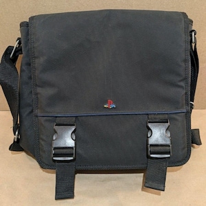 May include: A black messenger bag featuring a flap closure with two black plastic side-release buckles. The front of the bag displays a small, colorful PlayStation logo centered on the flap. The bag includes an adjustable black shoulder strap.