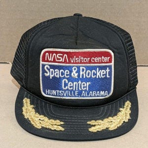 Puede incluir: Una gorra de camionero negra con un parche rectangular que muestra el texto NASA visitor center Space & Rocket Center HUNTSVILLE, ALABAMA. El parche tiene una sección superior roja y una inferior azul. Dos diseños de hojas de laurel doradas están bordados en la visera.