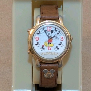Peut inclure: Une montre-bracelet à quartz Lorus avec un bracelet en cuir marron orné d'une icône Mickey Mouse. Le cadran blanc affiche Mickey Mouse, les chiffres rouges 3, 6, 9, 12 et de petites silhouettes de Mickey. Présentée sur un support en plastique beige.