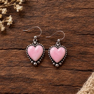 May include: A pair of drop earrings featuring smooth, light pink heart-shaped stones set in silver-toned metal. Each heart is framed by a decorative border of small silver beads, suspended from simple silver-toned metal ear wires for a classic look.