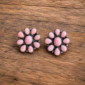 May include: A pair of stud earrings featuring a central oval pink stone surrounded by a ring of smaller teardrop-shaped pink stones set in oxidized silver. Each earring has a floral-inspired arrangement with a dark metal border and silver beads.