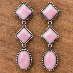 May include: A pair of drop earrings featuring three vertically aligned pink stones set in silver-toned metal. Each earring consists of two diamond-shaped stones at the top and one oval-shaped stone at the bottom, all framed by a beaded silver border.