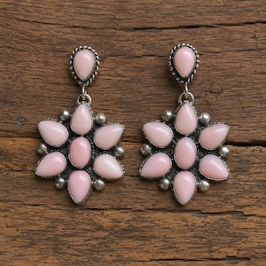 May include: A pair of silver drop earrings featuring a central oval pink stone surrounded by six teardrop-shaped pink stones arranged in a flower pattern. Each earring includes a small teardrop-shaped pink stone at the top with a twisted silver border.