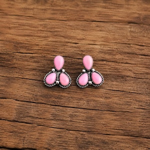 May include: A pair of stud earrings featuring three teardrop-shaped pink stones arranged in a cluster. Each stone is set in a silver-toned metal frame with a twisted rope texture and small silver beads between the stones, creating a floral shape.