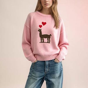 Puede incluir: Un suéter de punto de manga larga en color rosa claro con un gráfico de una llama marrón en el pecho. Sobre la llama hay dos pequeños corazones rojos pixelados. El suéter tiene cuello redondo, puños y dobladillo acanalados, combinado con jeans azules.