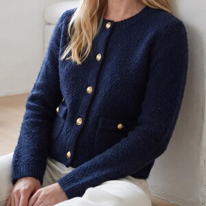 May include: A navy blue long-sleeved knit cardigan with a textured surface, a round neckline, and a front button closure featuring six polished gold-tone metal buttons. The garment includes two small front pockets with flaps and ribbed cuffs.