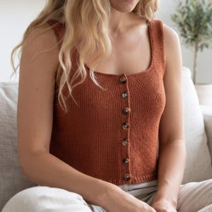 May include: A sleeveless, rust-colored knit tank top featuring a square neckline and a vertical front placket secured with seven round, brown, marbled buttons. The garment is constructed with a fine-gauge knit texture and a straight hemline.