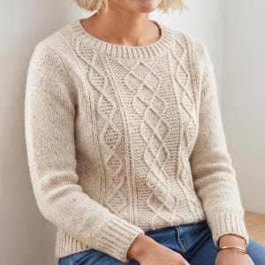 May include: A cream-colored long-sleeve sweater featuring a textured cable knit pattern with vertical diamond shapes and a ribbed crew neckline, cuffs, and hem. The knit fabric has subtle dark flecks throughout the light beige material.