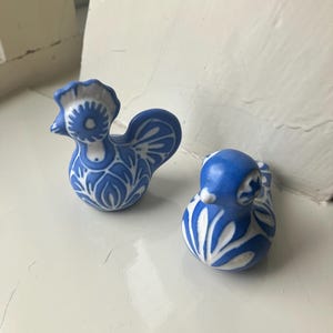 Puede incluir: Dos pequeñas figuras de pájaros de cerámica con un vibrante patrón floral azul y blanco. Una figura tiene forma de gallo con una cresta prominente, mientras que la otra es un pájaro redondeado con un cuerpo liso y curvo con motivos pintados.