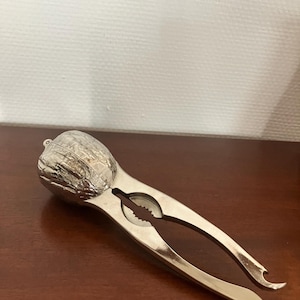 Op de afbeelding: Een zilverkleurige metalen notenkraker met een getextureerde, walnootvormige kop en twee langwerpige handvatten. Het gereedschap heeft een centraal gekarteld gedeelte voor het kraken van doppen en een klein haakje aan het uiteinde.