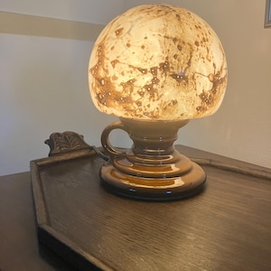 Op de afbeelding: Een tafellamp met een koepelvormige glazen kap met een gevlekt bruin en crèmekleurig patroon. De voet is gemaakt van glanzend bruin keramiek en heeft de vorm van een koffiemok met een handvat aan de zijkant, rustend op een getrapte, ronde voet.