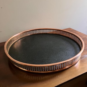 Op de afbeelding: Een rond serveerblad met een metalen, koperkleurige rand met een verticaal lattenontwerp. De basis van het blad is cirkelvormig en afgewerkt in een effen zwart materiaal met een getextureerd oppervlak, wat een contrast vormt met het koperen frame.