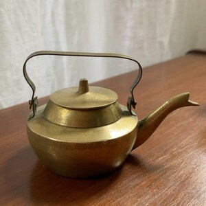 Op de afbeelding: Een kleine koperen theepot met een afgerond lichaam, een plat deksel met een centrale knop en een gebogen tuit. De theepot heeft een dun, gebogen metalen handvat aan de zijkanten en is afgewerkt met een goudkleurige, textuurrijke laag.