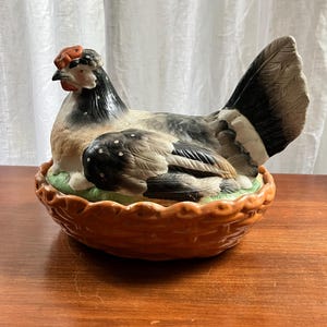 Può includere: Una statuetta di gallina in ceramica che riposa in un cestino color marrone in stile intrecciato. La gallina presenta un piumaggio nero, bianco e marrone chiaro con una cresta e bargigli rosso-arancio brillante. L'uccello poggia su una base verde.