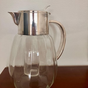 Op de afbeelding: Een glazen karaf met een geribbelde, ronde vorm en een deksel, tuit en handvat van zilverkleurig metaal. De karaf heeft een centrale, cilindervormige glazen inzet voor koeling, afgewerkt met een metalen deksel en een kleine hendel om te openen.