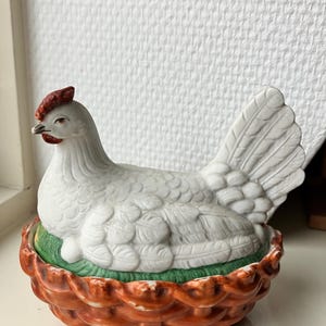 Op de afbeelding: Een keramieken henfiguur rustend in een basis met vlechtwerkpatroon. De hen is wit met een rode kam en lellen. De basis is beschilderd in glanzend oranje-bruin om een mand na te bootsen, met een stukje groen gras onder de hen.