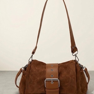 May include: A brown suede shoulder bag featuring a smooth brown leather strap and a central front flap secured by a silver-toned metal buckle. The bag includes a matching brown leather shoulder strap attached with silver-toned metal hardware clips.