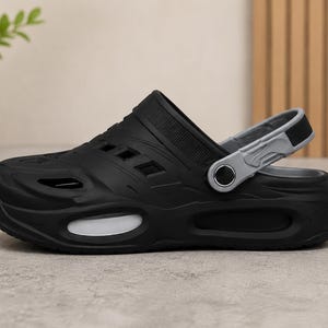 May include: A black clog shoe featuring a thick sole with white oval side accents, multiple ventilation cutouts across the upper, and a pivoting gray heel strap attached with circular silver-toned hardware on the side of the footwear.