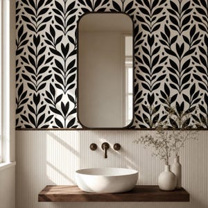 Puede incluir: Un área de lavabo de baño con un lavabo blanco sobre un estante de madera, un grifo de pared y un espejo rectangular con esquinas redondeadas. La pared tiene papel tapiz con patrón de hojas en blanco y negro sobre paneles verticales blancos.